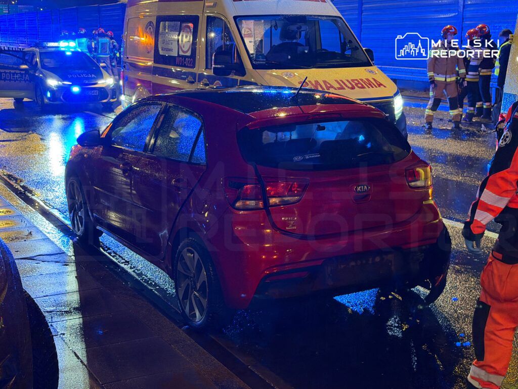 Czerwony samochód osobowy zaparkowany obok karetki pogotowia i radiowozu policyjnego na mokrej nawierzchni.