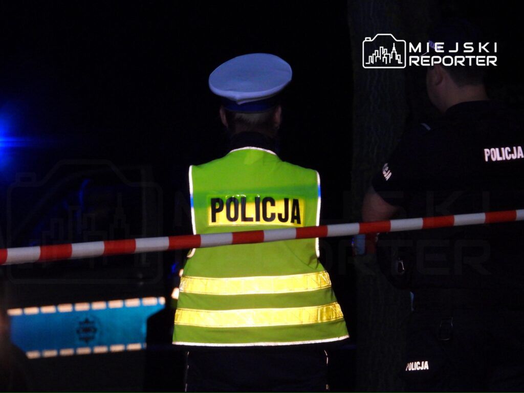 Policjanci na miejscu zdarzenia, zabezpieczając teren policyjnym taśmą. W tle widać niebieskie światła radiowozu. Sytuacja ma miejsce w nocy, co sugeruje pilny charakter interwencji. Funkcjonariusze w odblaskowych kamizelkach monitorują sytuację.