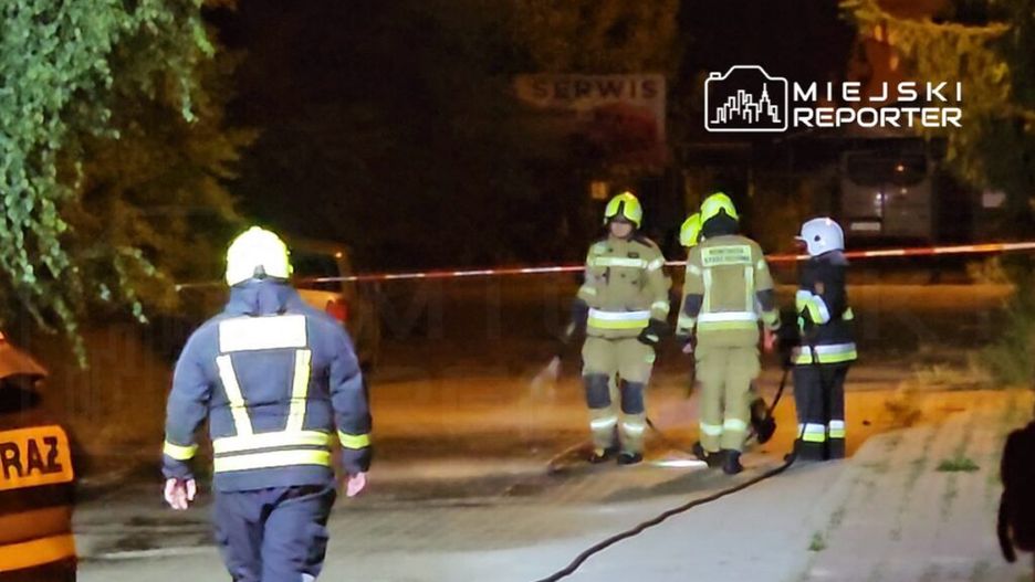 Grupa strażaków w mundurach gaśniczych prowadzi działania na oświetlonej ulicy, przy taśmie ostrzegawczej.