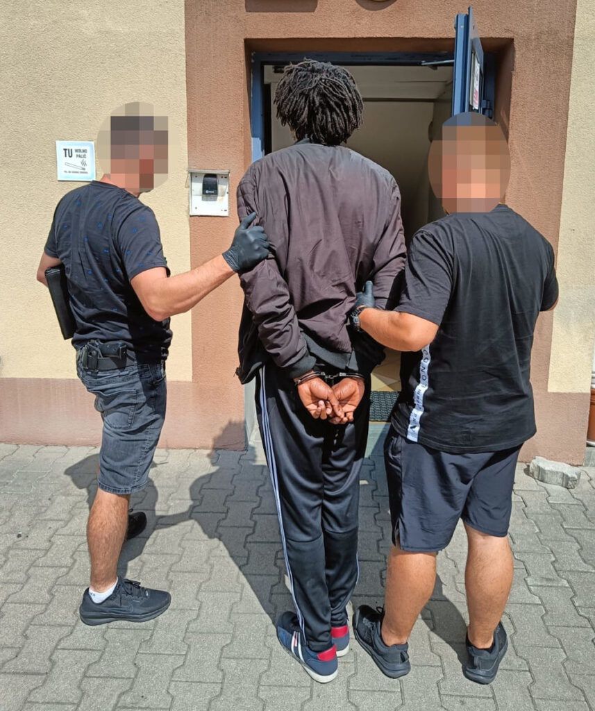 Mężczyzna w dresie prowadzony przez dwóch funkcjonariuszy policji w kierunku budynku.