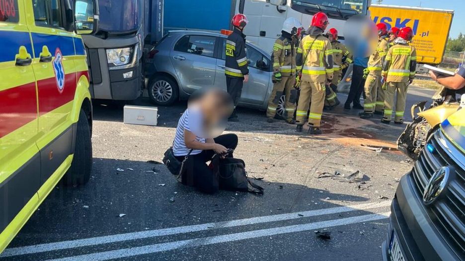 Kobieta w pasiastym T-shircie siedzi na jezdni obok wraku samochodu, podczas gdy strażacy udzielają pomocy poszkodowanym.