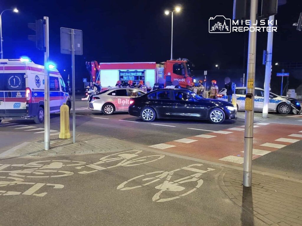 Na skrzyżowaniu zatrzymane pojazdy, w tym karetka i radiowóz, podczas nocnej interwencji służb.
