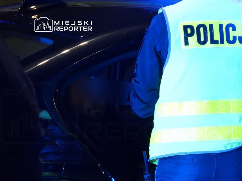 Policjant w odblaskowej kamizelce kontroluje wnętrze samochodu w nocy, w otoczeniu niebieskich świateł.