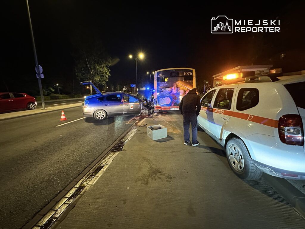 Samochód osobowy z uszkodzeniami stoi na drodze, obok interweniujący funkcjonariusz w pobliżu autobusu z numerem 1076.
