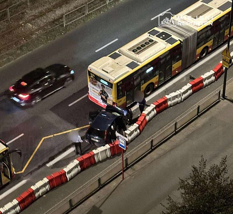 Czarny samochód zatrzymany przy barierkach, obok przystanku autobusowego, w nocy, obok pasażerów wysiadających z autobusu.
