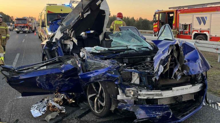 Rozbity niebieski samochód osobowy z uszkodzoną karoserią leży na drodze, obok stoją strażacy i karetka.
