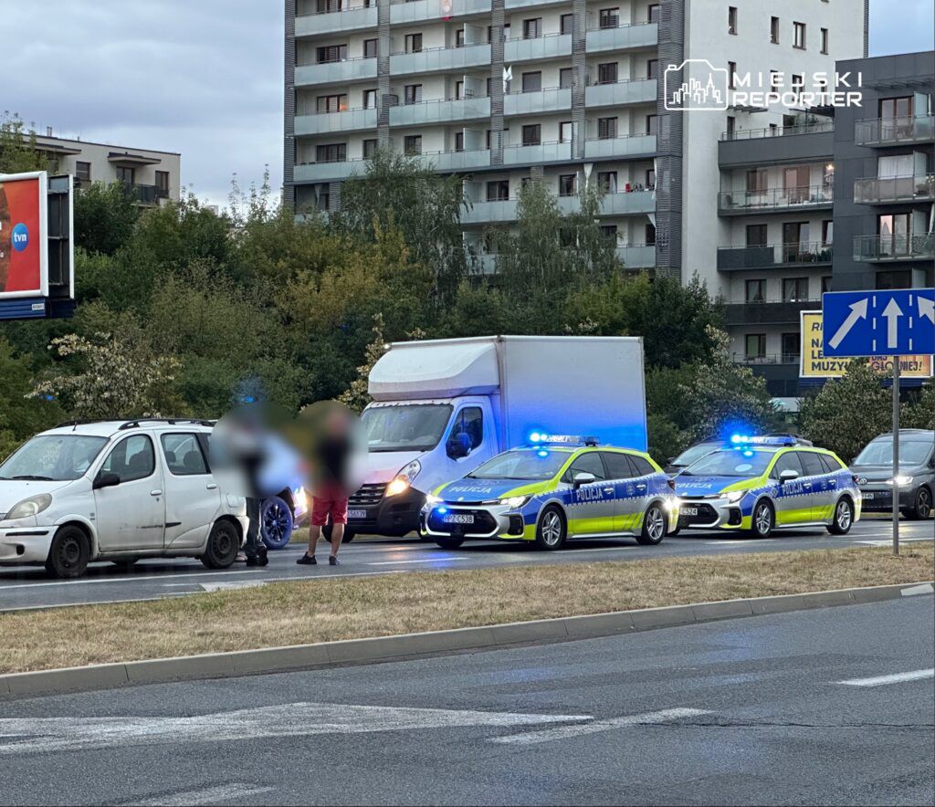 Na drodze zatrzymane dwa radiowozy policyjne z włączonymi sygnałami świetlnymi, obok stoi mężczyzna rozmawiający z kierowcą białego samochodu.