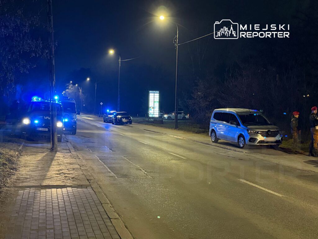 Na oświetlonej ulicy nocą zaparkowane są pojazdy policyjne z włączonymi sygnałami świetlnymi, a obok stoją funkcjonariusze.