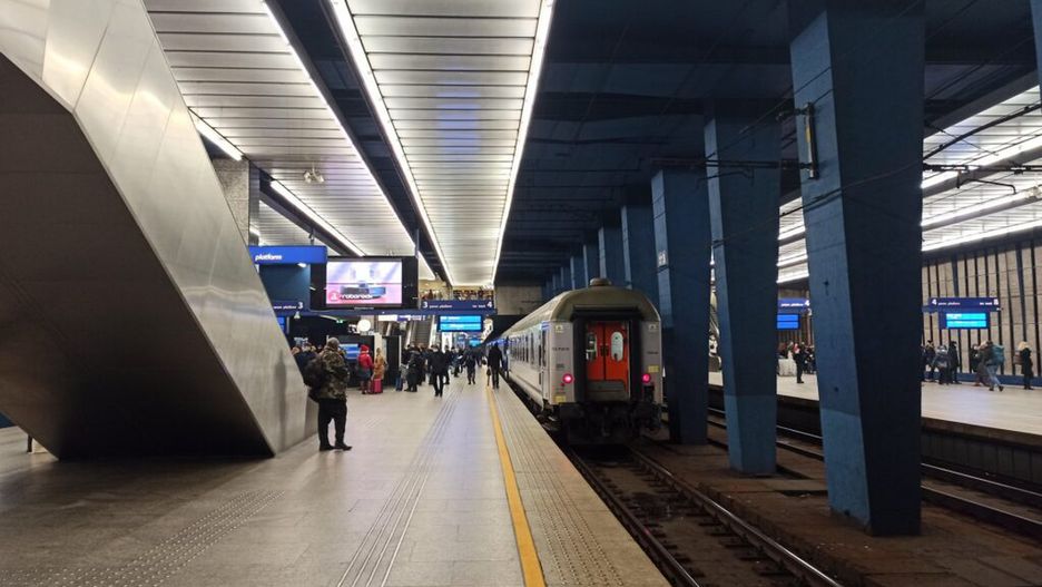 Pasażerowie czekają na peronie stacji kolejowej, obok zatrzymany pociąg w nowoczesnym wnętrzu z niebieskimi filarami.