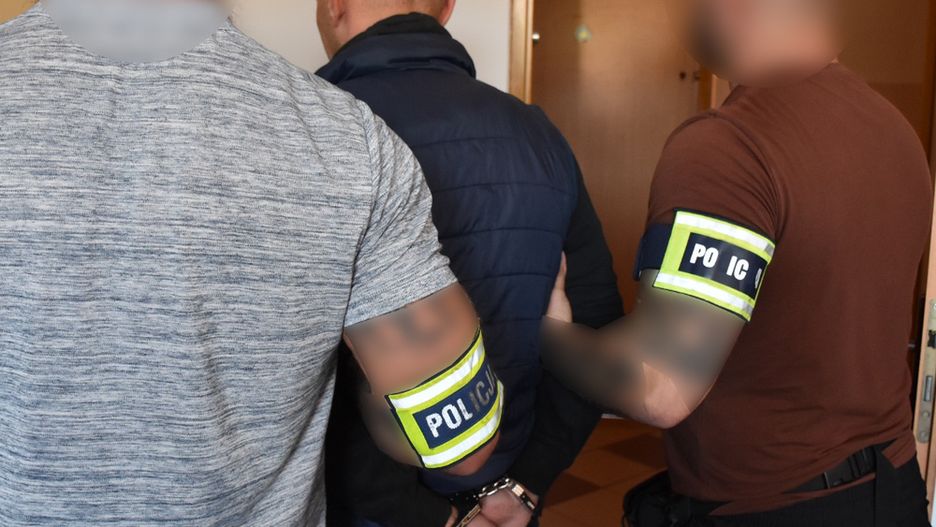 Trzej policjanci w mundurach z odznakami prowadzą mężczyznę w kajdankach w korytarzu budynku.