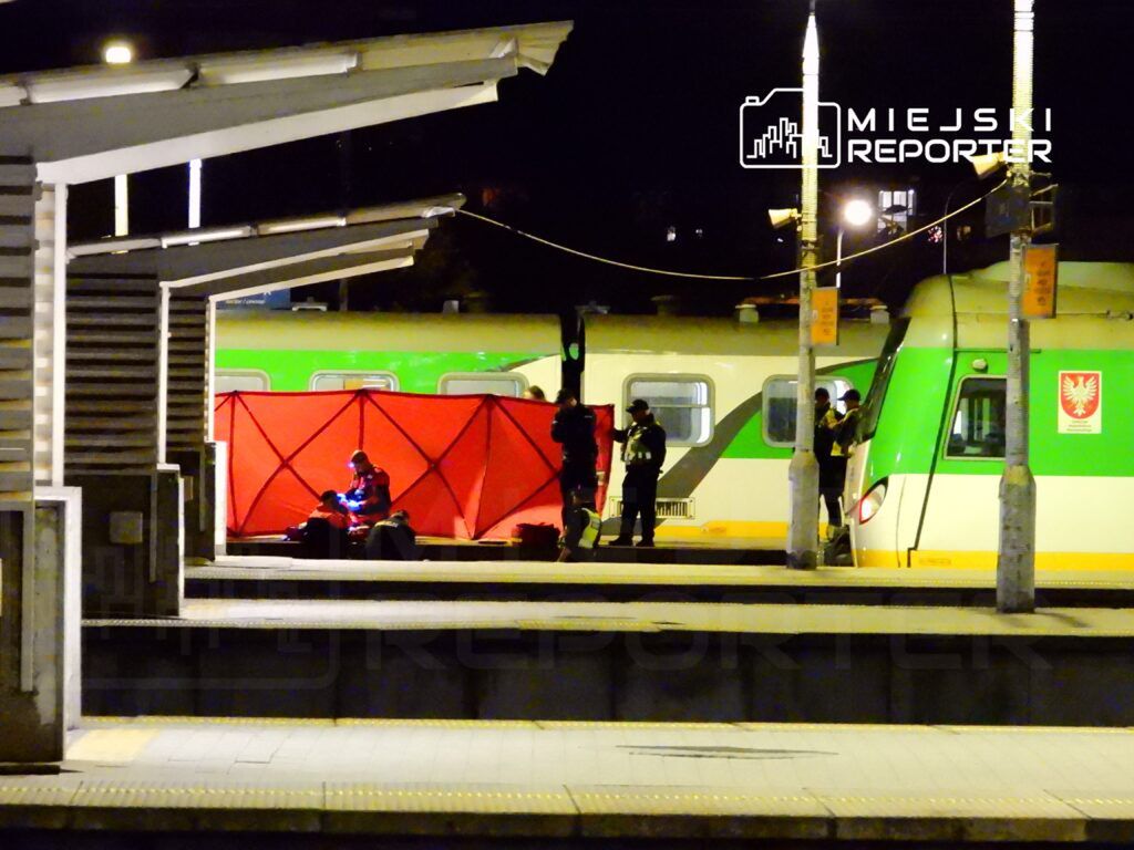 Ratownicy w mundurach pracują przy czerwonym namiocie na stacji kolejowej, obok pociągu w nocy.