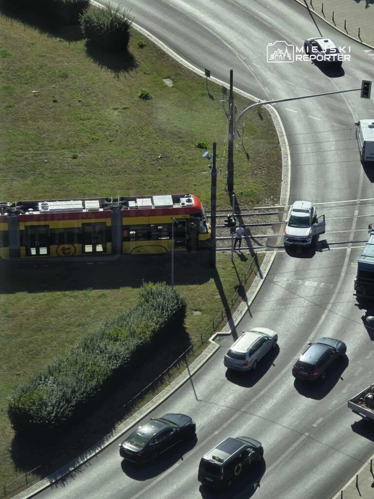 Tramwaj z czerwoną i żółtą obudową zatrzymany na przejeździe, obok kręta ulica z ruchem samochodowym.