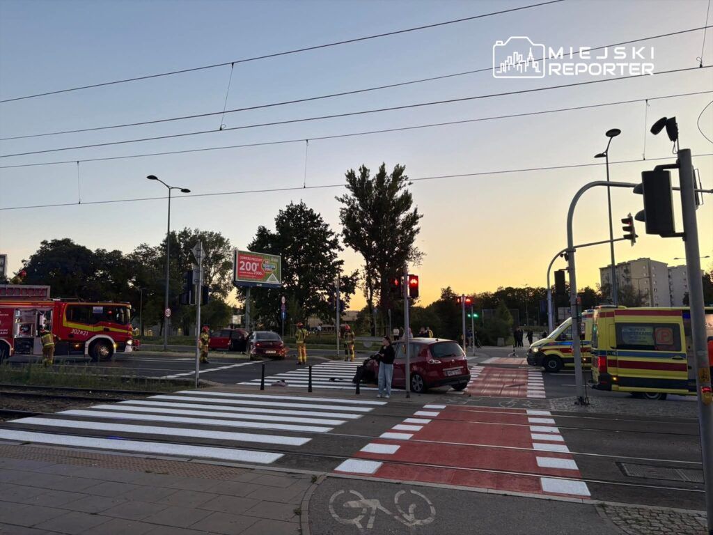 W miejscu zdarzenia drogowego, strażacy i ratownicy medyczni pracują przy uszkodzonym samochodzie na przejściu dla pieszych.