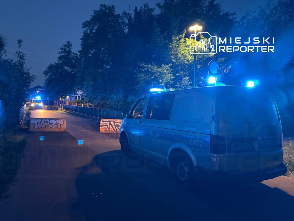 Policyjny van z niebieskimi światłami zatrzymany przy barierkach z graffiti na drodze w nocy.