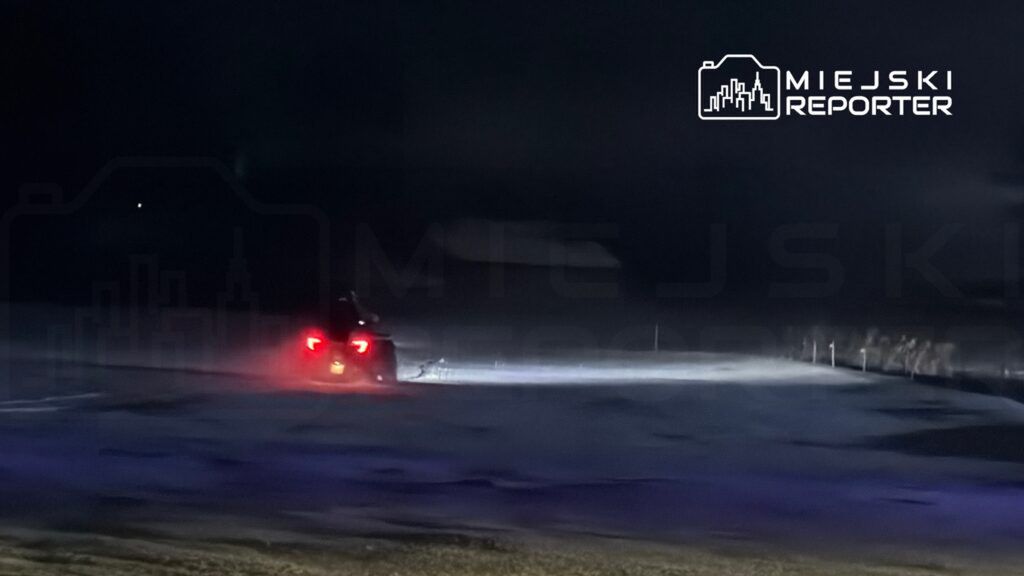 Samochód porusza się po zaśnieżonej drodze w ciemności, emitując jasne światło przednie.