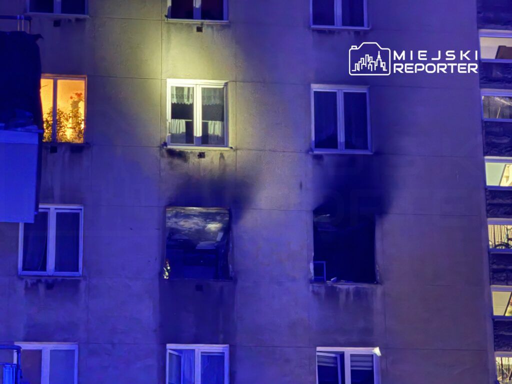 Na elewacji budynku widać okna, z których jedno jest oświetlone, a obok znajduje się okno z uszkodzeniami po pożarze.