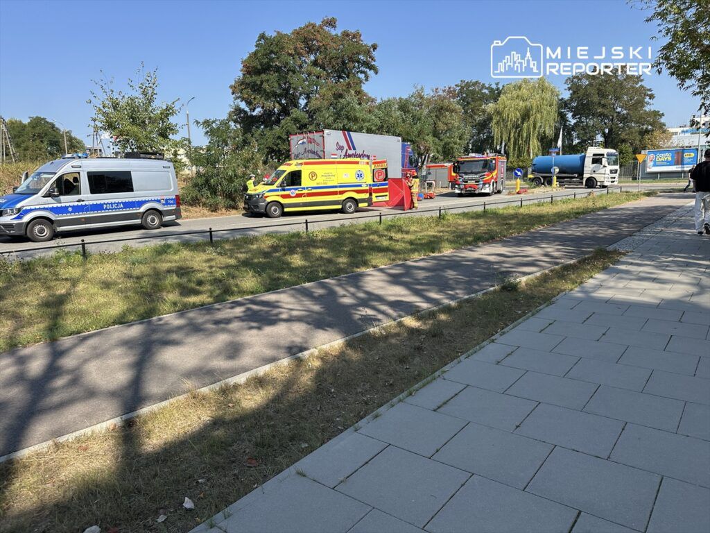 Na poboczu ulicy zaparkowane są pojazdy służb ratunkowych: radiowóz policji, karetka i wóz strażacki.