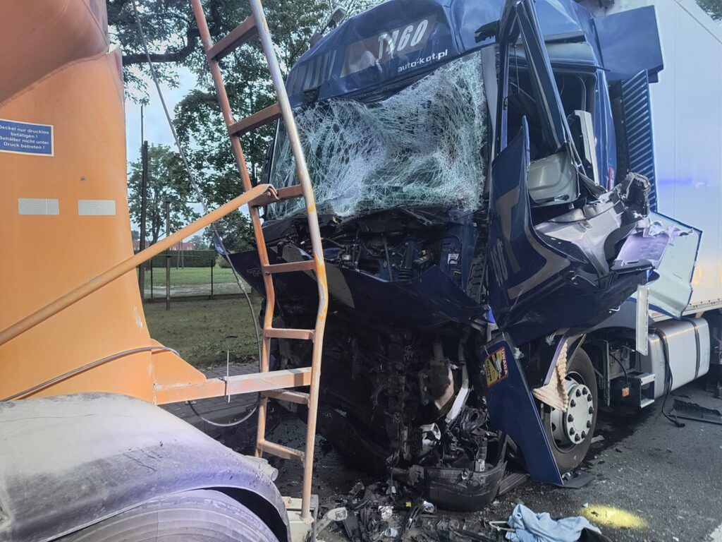 Wysokotonażowy pojazd ciężarowy z uszkodzoną kabiną i rozbitym przednim szkłem stoi obok innej ciężarówki na drodze.