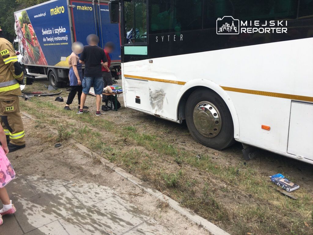 Grupa osób stoi obok uszkodzonego autobusu, a obok nich pracują strażacy w mundurach.