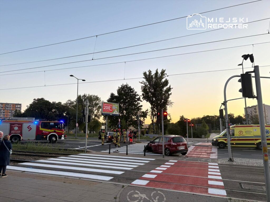 Na przejściu dla pieszych stoi czerwona osobówka z uszkodzoną stroną, obok pracują strażacy i ratownicy medyczni.