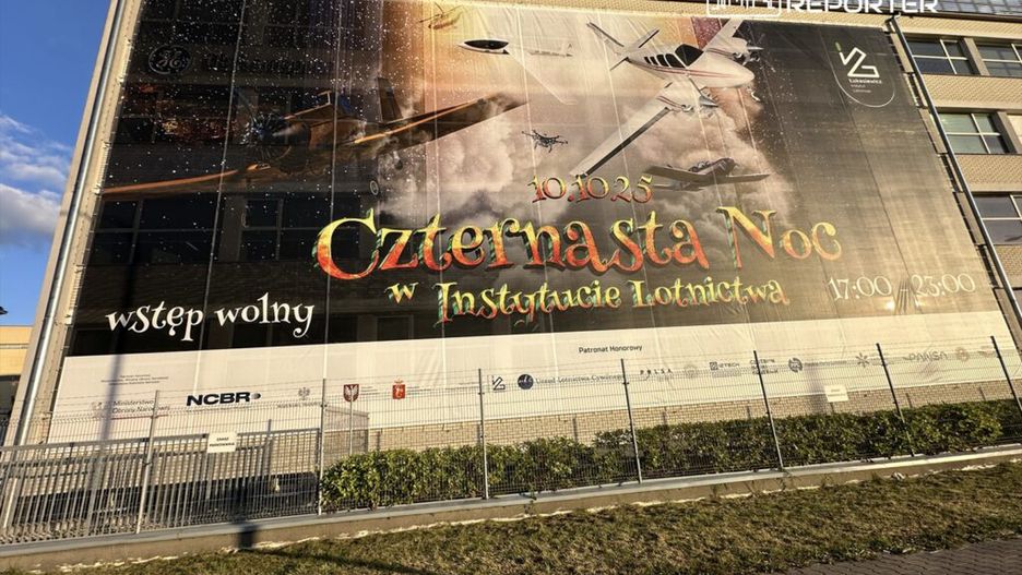 Billboard promujący wydarzenie "Czternasta Noc w Instytucie Lotnictwa" z datą i godzinami, otoczony zielenią.