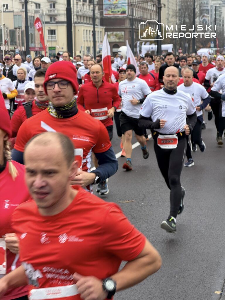 Grupa biegaczy w czerwonych i białych koszulkach uczestniczy w biegu ulicznym na miejskiej ulicy.