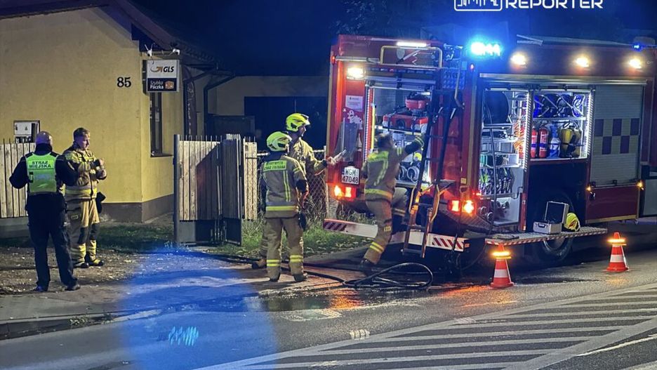 Strażacy w hełmach i mundurach pracują przy wozie strażackim na ulicy, obok budynku z oznaczeniem GLS.