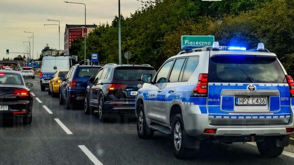 Policyjny samochód terenowy z włączonymi sygnałami świetlnymi stoi w korku na drodze. W tle widoczny znak kierunkowy do Piaseczna.