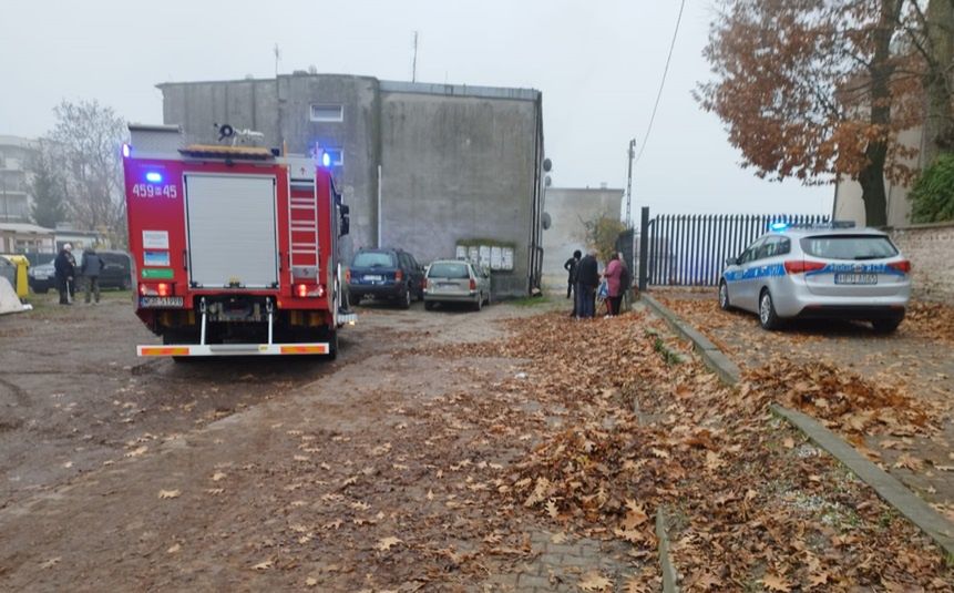 W tle szarego budynku, straż pożarna z włączonymi sygnałami świetlnymi, obok policyjny radiowóz, a w pobliżu grupka ludzi rozmawia.
