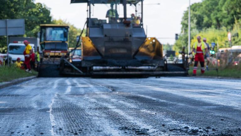Maszyna budowlana asfaltująca drogę, obok pracownicy w odzieży roboczej, w tle zieleń i znaki drogowe.