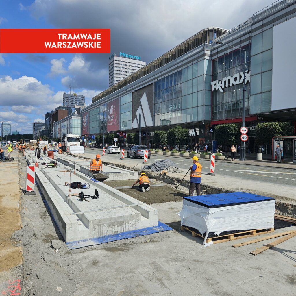 Grupa pracowników w pomarańczowych kamizelkach roboczych układa tory tramwajowe na miejskiej ulicy, obok nowoczesnych budynków.