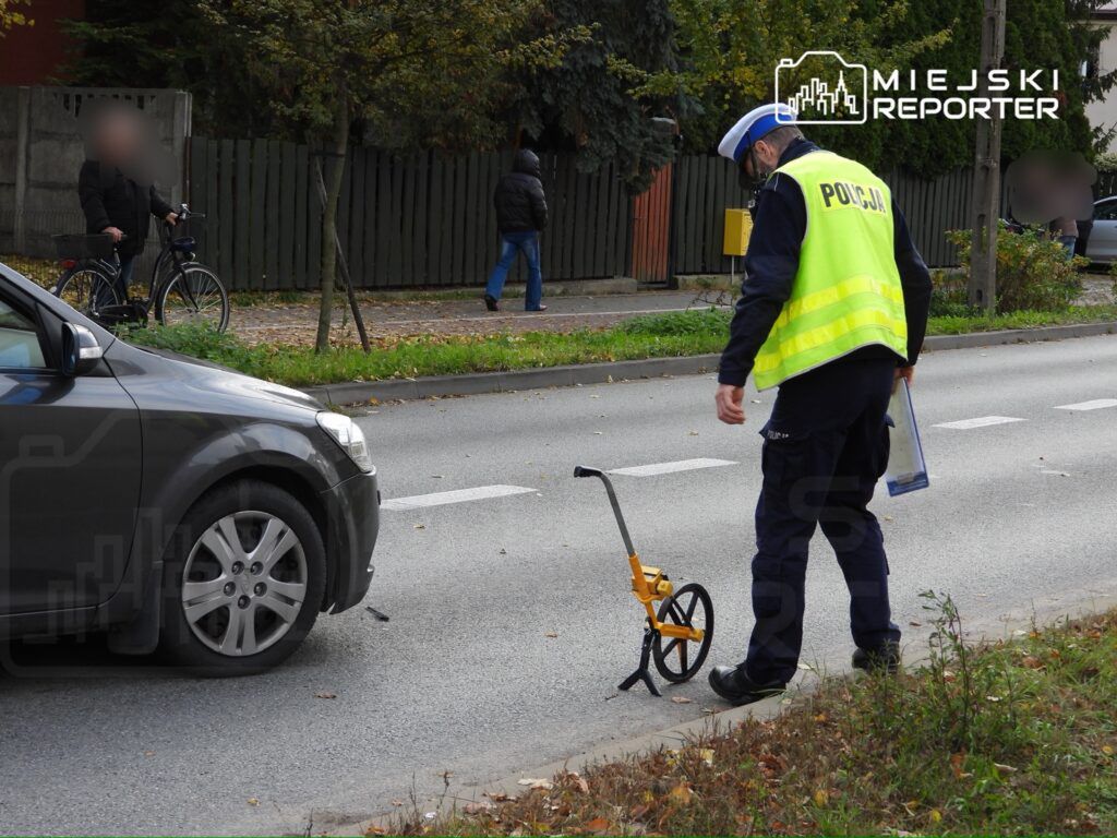 Policjant w kamizelce odblaskowej mierzy drogę przy zaparkowanym samochodzie na ulicy.