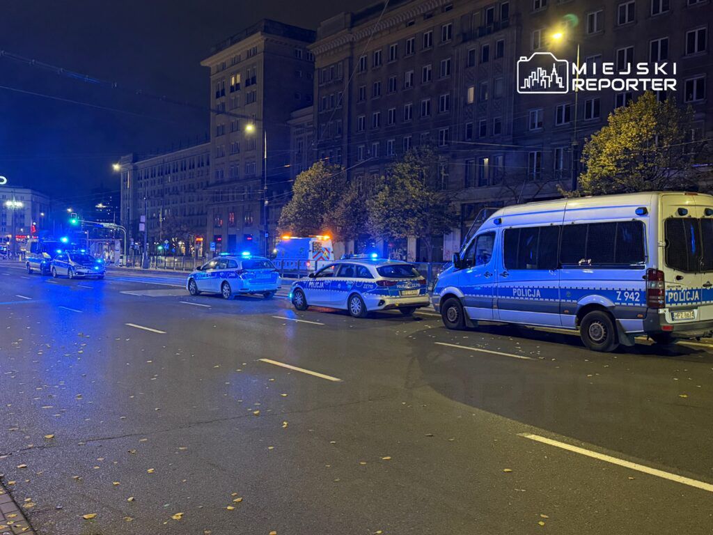 Policyjne pojazdy z włączonymi sygnałami świetlnymi blokują ulicę w mieście nocą, w tle widoczne oświetlone budynki.