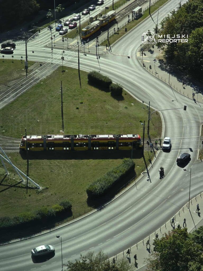 Tramwaj w kolorze żółtym przejeżdża przez skrzyżowanie, obok ruchliwa ulica z samochodami i pieszymi.