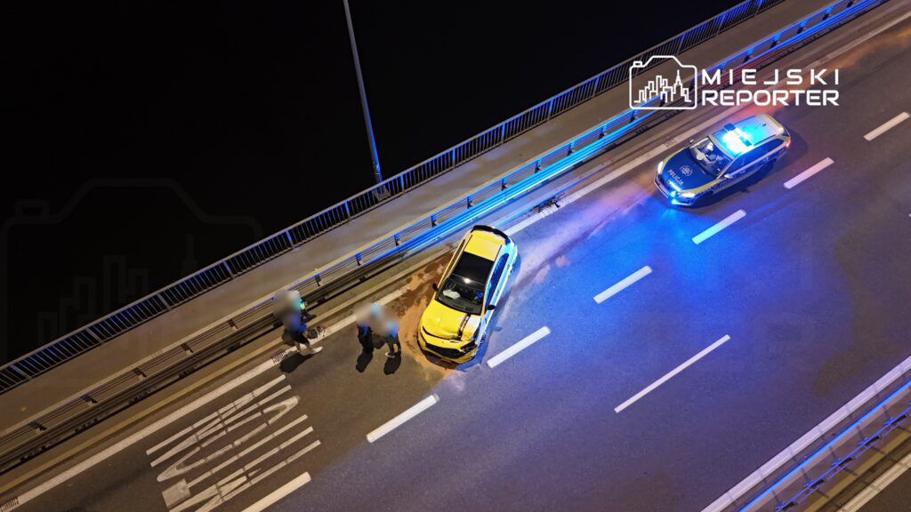 Na drodze stoi uszkodzony żółty samochód, obok dwóch osób rozmawia z policjantami przy radiowozie z włączonymi sygnałami.