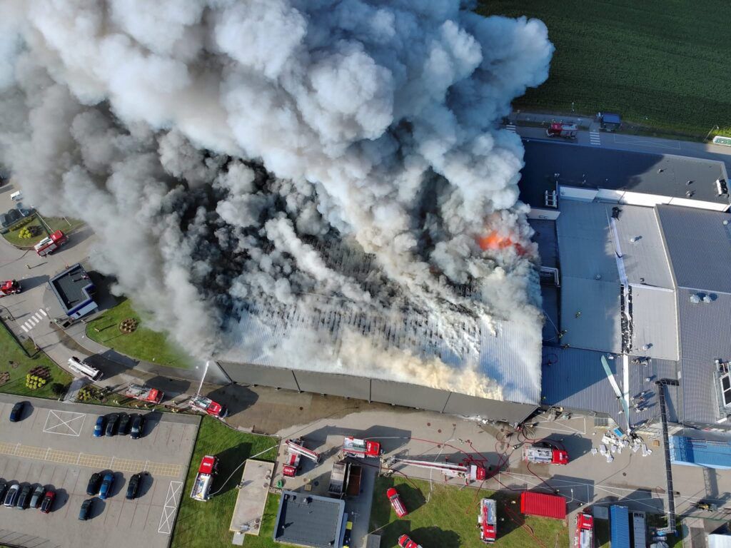 Gęsty dym i ogień wydobywają się z dachu dużego budynku, wokół którego pracują strażacy z wężami gaśniczymi.