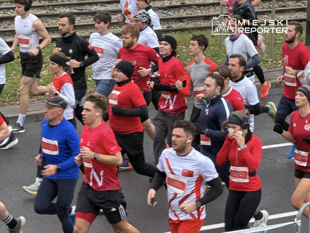 Grupa biegaczy w czerwonych i białych koszulkach uczestniczy w wyścigu na ulicy, obok torów kolejowych.
