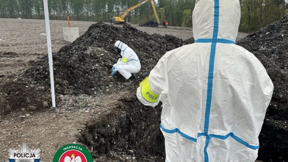 Funkcjonariusze w kombinezonach ochronnych przeszukują teren z odpadami, w tle widoczny koparka.