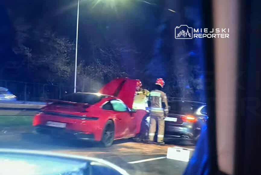 Czerwony samochód sportowy z otwartą maską, przy którym stoi strażak w hełmie, na drodze w nocy.