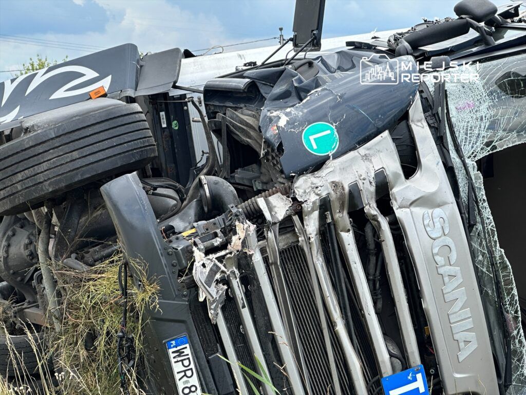 Przewrócony ciężarówka marki Scania leży na boku w zaroślach, z uszkodzoną kabiną i rozbitą szybą.