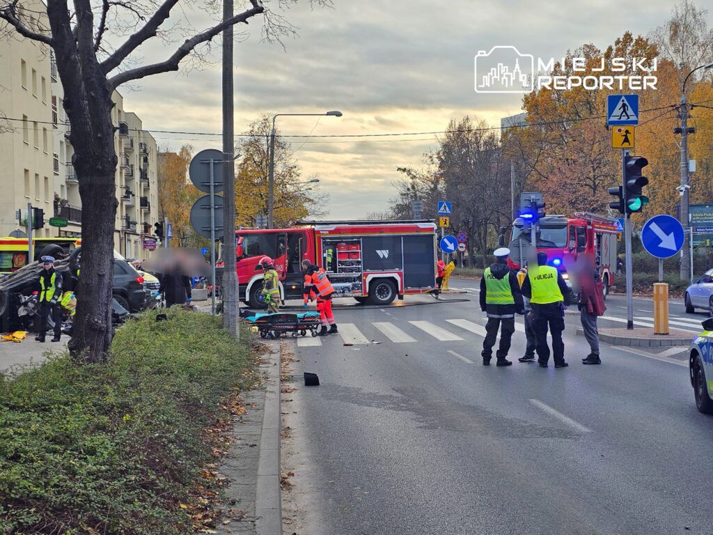 Na skrzyżowaniu ulicy interweniują strażacy i policjanci, zablokowane są pojazdy. W tle widoczny wóz strażacki.