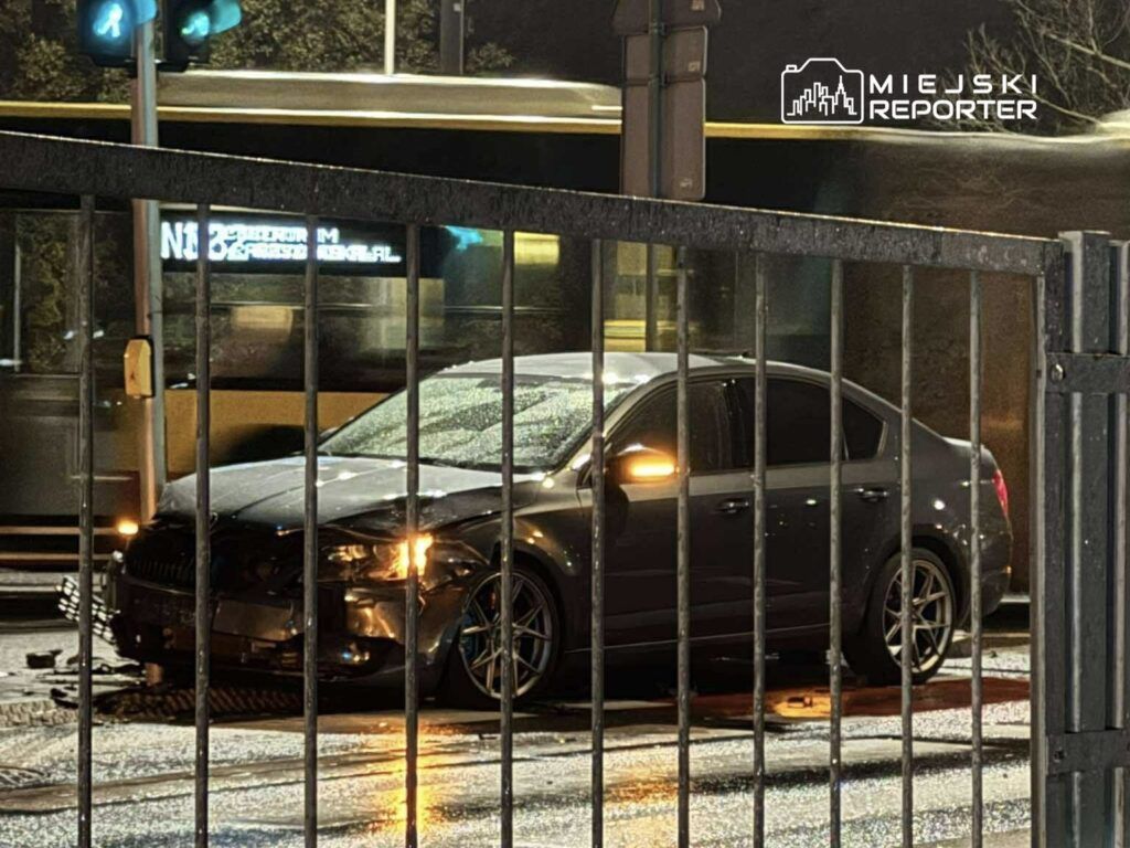 Uszkodzony samochód osobowy stoi na mokrej nawierzchni w nocy, za ogrodzeniem, obok przejeżdżającego tramwaju.