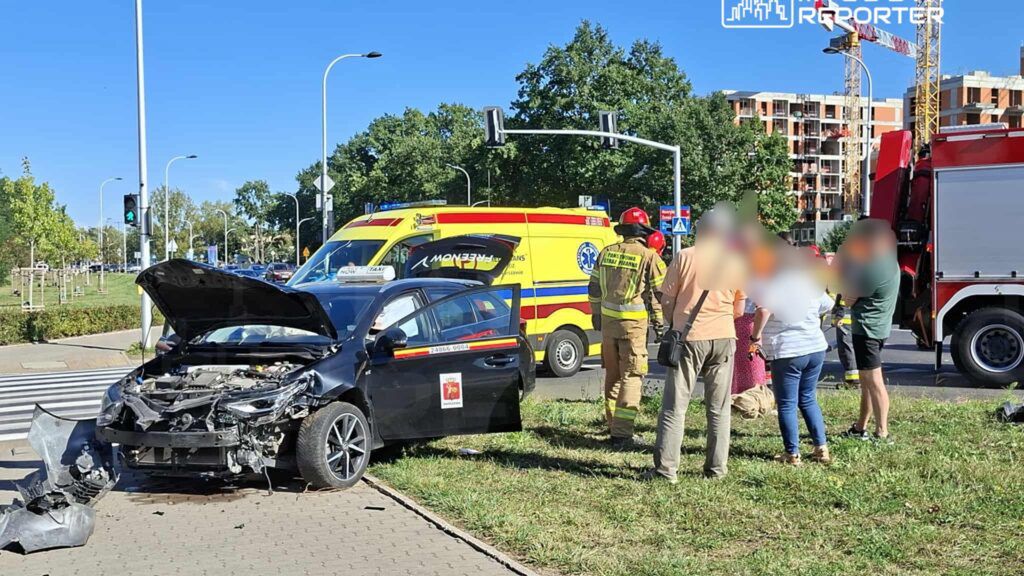Czarny samochód osobowy z uszkodzoną maską stoi na trawie obok jezdni, wokół niego grupa osób oraz służby ratunkowe.