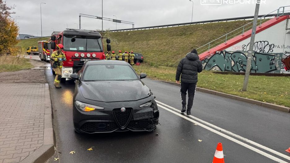 Kierowca w czarnej limuzynie z uszkodzeniem zjeżdża na pobocze, obok strażacy w akcji przy wozie strażackim.