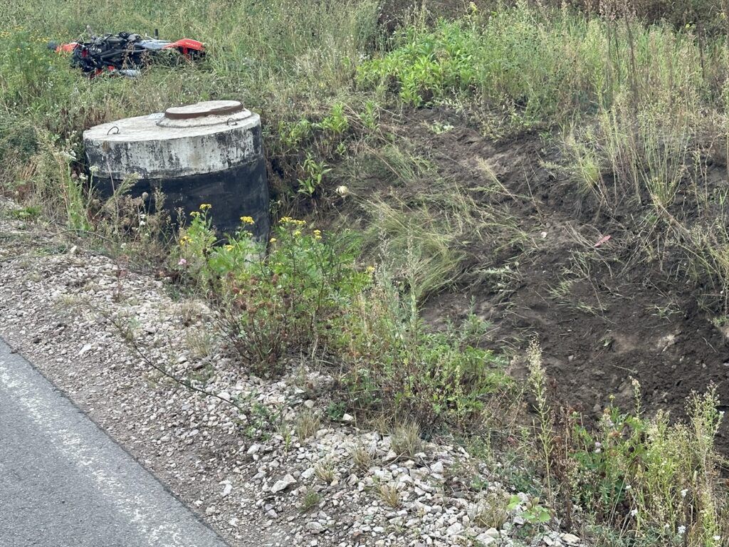 Betonowy zbiornik z metalową pokrywą w otoczeniu wysokiej trawy i krzewów, obok leży przewrócony motocykl.