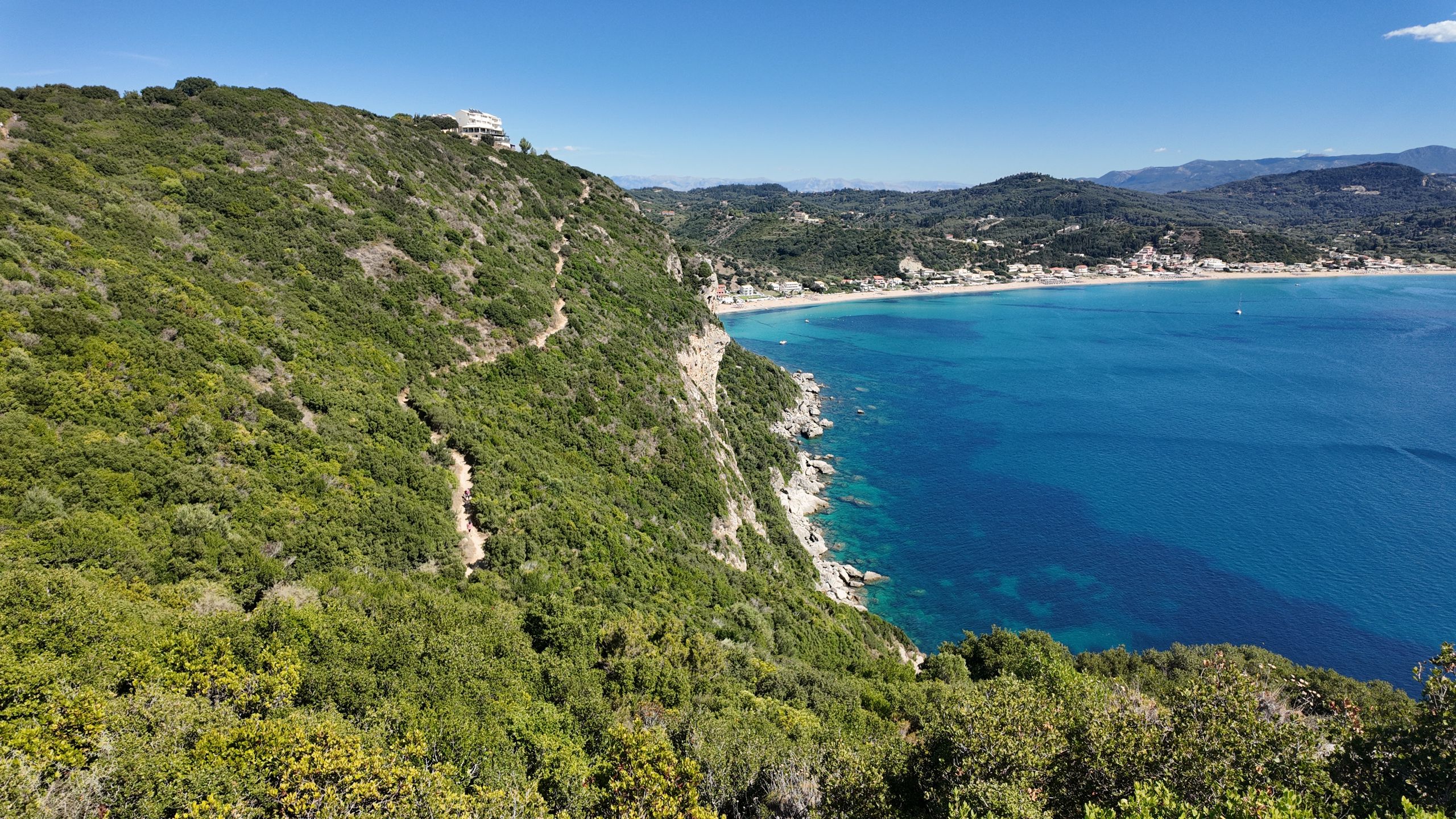 Ścieżka na zielonym, skalistym zboczu poniżej Afionas, a w tle turkusowa zatoka i długa plaża Agios Georgios Pagon na Korfu.