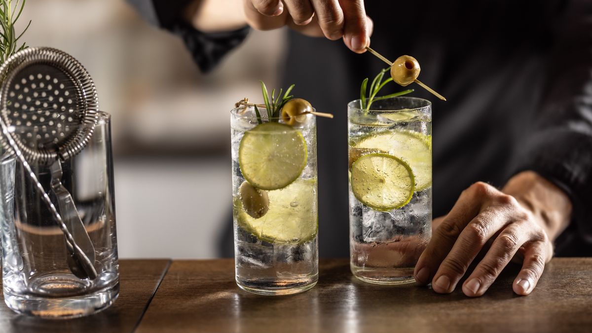 A close-up view of two Gin Tonic cocktails in tall glasses filled with ice and lime. The bartender's hands garnish both drinks with rosemary and an olive on a skewer.
A close-up view of two Gin Tonic cocktails in tall glasses filled with ice and lime. The bartender's hands garnish both drinks with rosemary and an olive on a skewer.
SimpleImages