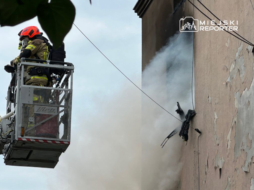 Strażacy w podnośniku gaszą pożar na ścianie budynku, z którego wydobywa się dym.