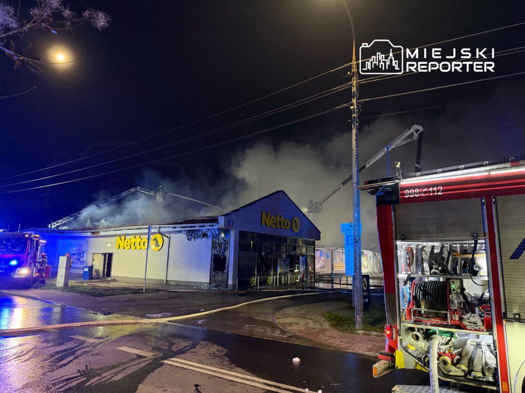 Pożar w sklepie Netto, z którego wydobywa się dym; strażacy używają drabiny do gaszenia ognia.