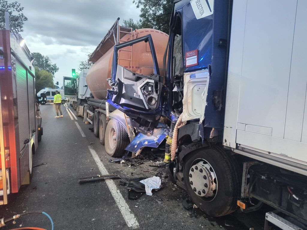 Wypadek drogowy z udziałem dwóch ciężarówek, uszkodzona kabina jednej z nich, w tle strażacy i policja na miejscu zdarzenia.